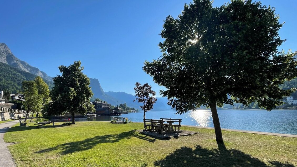 <p>Der Badeplatz Holzplatz/Seeklause direkt am Wasser vom wunderschlnen Grundlsee ist von grünen Wiesen und Bäumen umgeben. Im Hintergrund sind die majestätischen Berge und ein klarer blauer Himmel zu sehen.</p> | © Tourismusverband Ausseerland