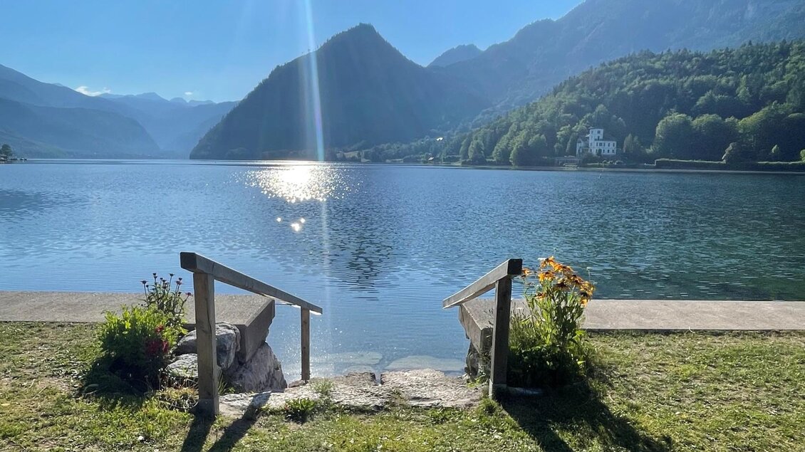 <p>Der ruhige Grundlsee mit glitzerndem Wasser und den umliegenden Bergen des Ausseerlandes. Im Vordergrund ist der Seezugang mit Blumen zu sehen.</p> | © Tourismusverband Ausseerland