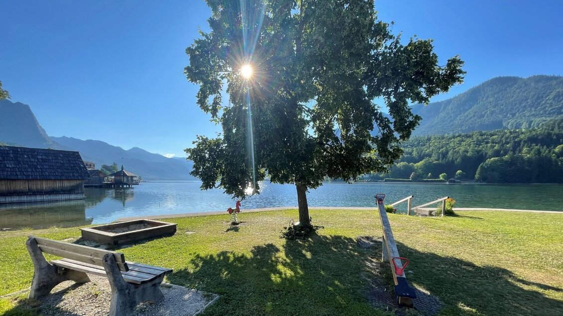 Der ruhige Grundlsee mit den majestätischen Bergen vom Ausseerland im Hintergrund. Ein Baum spendet Schatten, während die Sonne kräftig strahlt. | © Tourismusverband Ausseerland