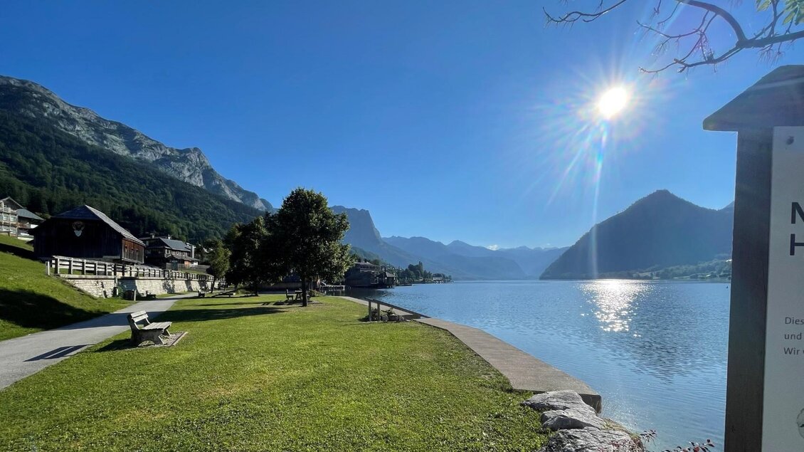 Das ruhige Seeufer vom Grundlsee mit den majestätischen Bergen vom Ausseerland im Hintergrund und strahlendem Sonnenschein. Die sanfte Landschaft ist von sanften grünem Gras und Bäumen umgeben. | © Tourismusverband Ausseerland