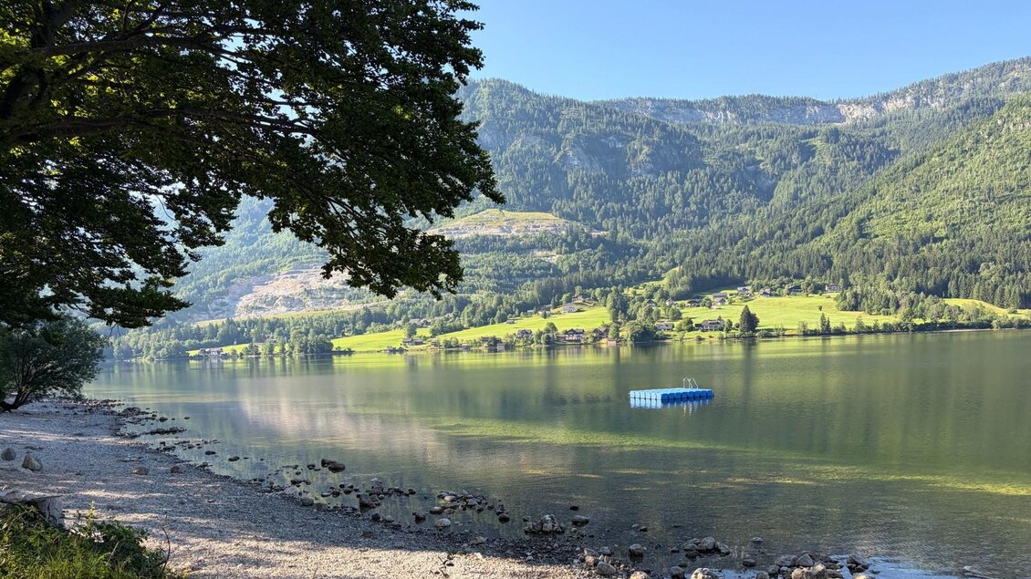 Der ruhige Grundlsee umgeben von majestätischen Bergen und saftig grünen Bäumen. Im Wasser schwimmt ein blaues Floß. | © Tourismusverband Ausseerland