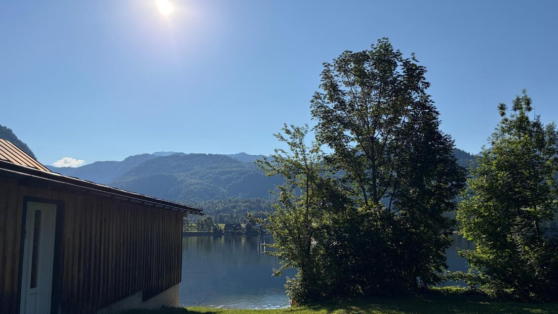 <p>Der ruhige Grundlsee mit den majestätischen Bergen im Hintergrund und einem klarem Himmel. Am Ufer steht ein Bootshaus und daneben grüne Bäume.</p> | © Tourismusverband Ausseerland