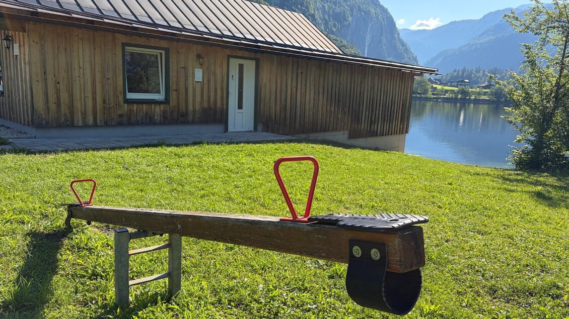 Eine Wippe steht auf einer saftig grünen Wiese vor einem Bootshaus. Im Hintergrund sind die Berge von Grundlsee und ein blauer Himmel zu sehen. | © Tourismusverband Ausseerland