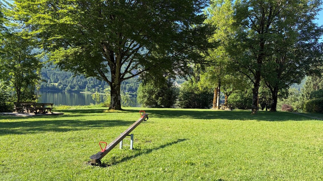 <p>Der grüne Badeplatz bei der Schachen-Siedlung mit einer Wippe und hohen Bäumen im Hintergrund. Dahinter ist der Grundlsee sichtbar.</p> | © Tourismusverband Ausseerland