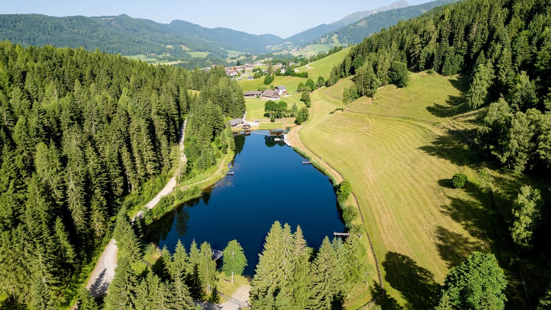 Ein ruhiger See umgeben von grünen Wäldern und sanften Hügeln. Im Hintergrund sind kleine Häuser und die Berge zu sehen. | © TV Murau