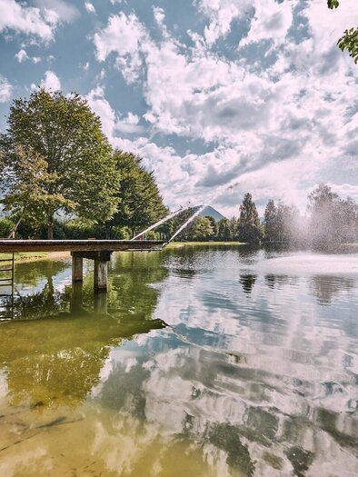 Ein ruhiger See umgeben von grünen Bäumen und sanften Hügeln. Der Himmel ist klar mit einigen weißen Wolken. | © Thomas Sattler