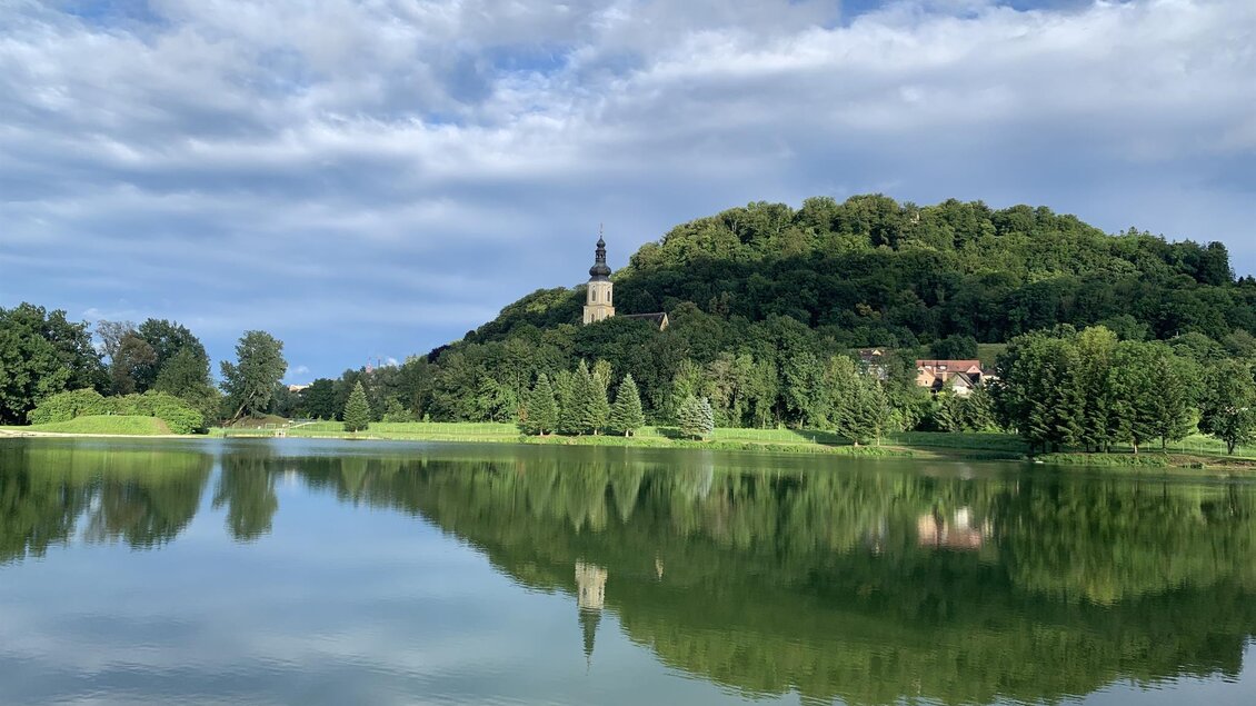 Ein ruhiger See spiegelt die grüne Hügel und den Himmel wider. Im Hintergrund erhebt sich ein Aussichtsturm auf dem Hügel. | © Lisi Maier