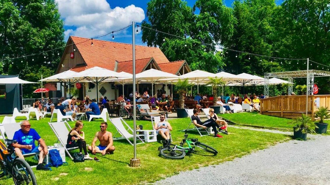 Ein belebtes Café im Freien mit vielen Sitzplätzen und Sonnenschirmen. Gäste entspannen auf Liegen und genießen die sonnige Atmosphäre. | © Wildon am See