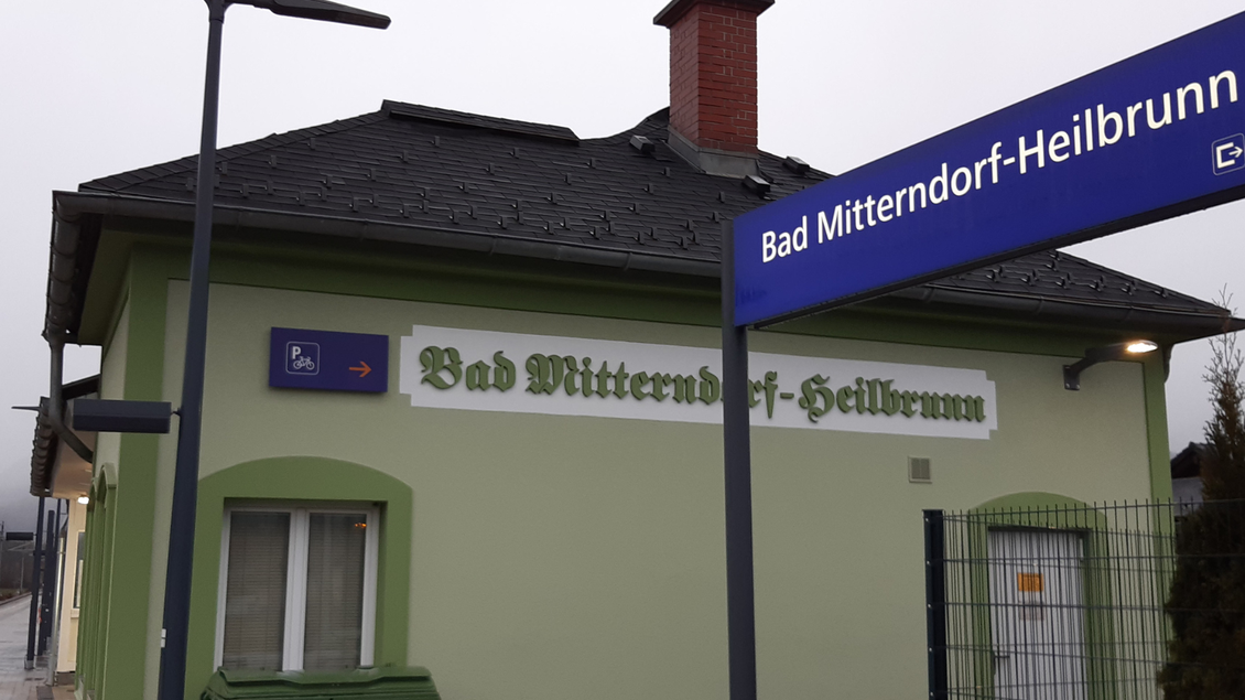 Ein Bahnhofsgebäude in Bad Mitterndorf-Heilbrunn mit grünem Anstrich und einem schrägen Dach. Der Bahnhofsschild zeigt den Namen des Ortes und einige Informationen. | © TVB Ausseerland Salzkammergut_Daniela Casari