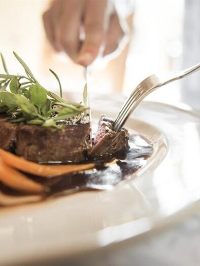 Ein köstliches Gericht mit zartem Steak, Gemüse und einer leckeren Sauce. Eine Hand schneidet das Fleisch auf einem eleganten Teller. | ©  Ballonhotel Thaller