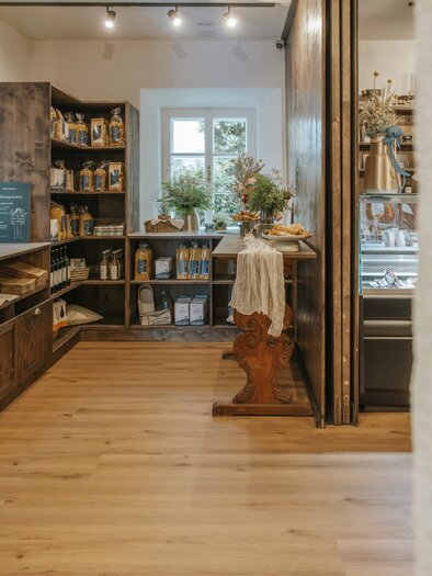 A cozy shop with shelves full of groceries and a refrigerated display case. The floor is made of wood and there is a decorative table arrangement.