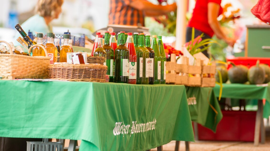 Ein Marktstand mit grünen Tischen, auf denen Flaschen und Körbe stehen. Im Hintergrund sind weitere Verkaufsstände und Menschen zu sehen. | © Tourismusverband Oststeiermark