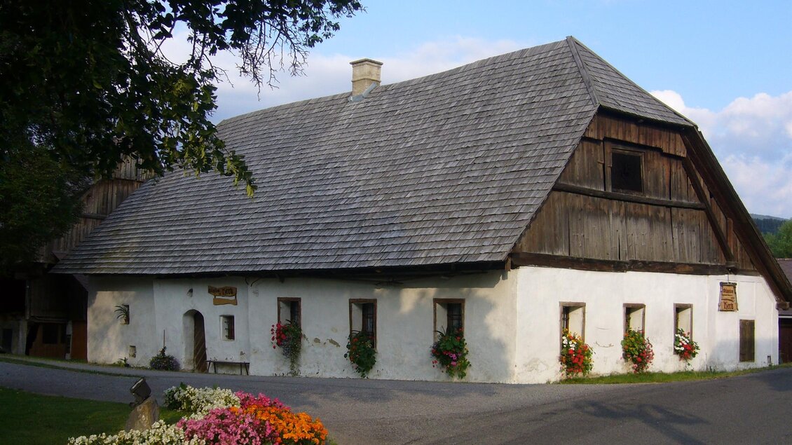 Ein traditionelles Bauernhaus mit einem steilen Dach und Holzverkleidung. Vor dem Haus wachsen bunte Blumen und es gibt eine ruhige Umgebung. | © Bauernmuseum Herkhof