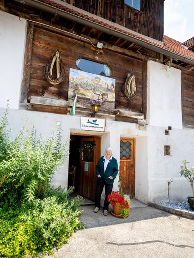 Ein historisches Gebäude mit einer ansprechenden Fassade aus Holz und Naturstein. Vor der Tür steht eine Person mit Blumen, umgeben von grünen Pflanzen. | © TVB Murau
