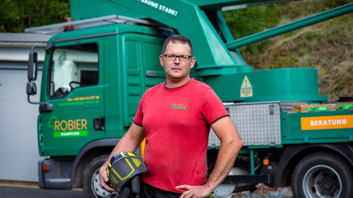 Ein Mann trägt einen Schutzhelm und steht vor einem grünen Dienstfahrzeug. Im Hintergrund ist eine Baustelle zu sehen. | © Baumpflege Robier