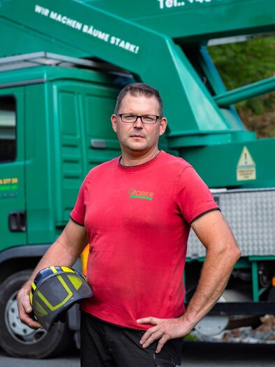 Ein Mann trägt einen Schutzhelm und steht vor einem grünen Dienstfahrzeug. Im Hintergrund ist eine Baustelle zu sehen. | © Baumpflege Robier