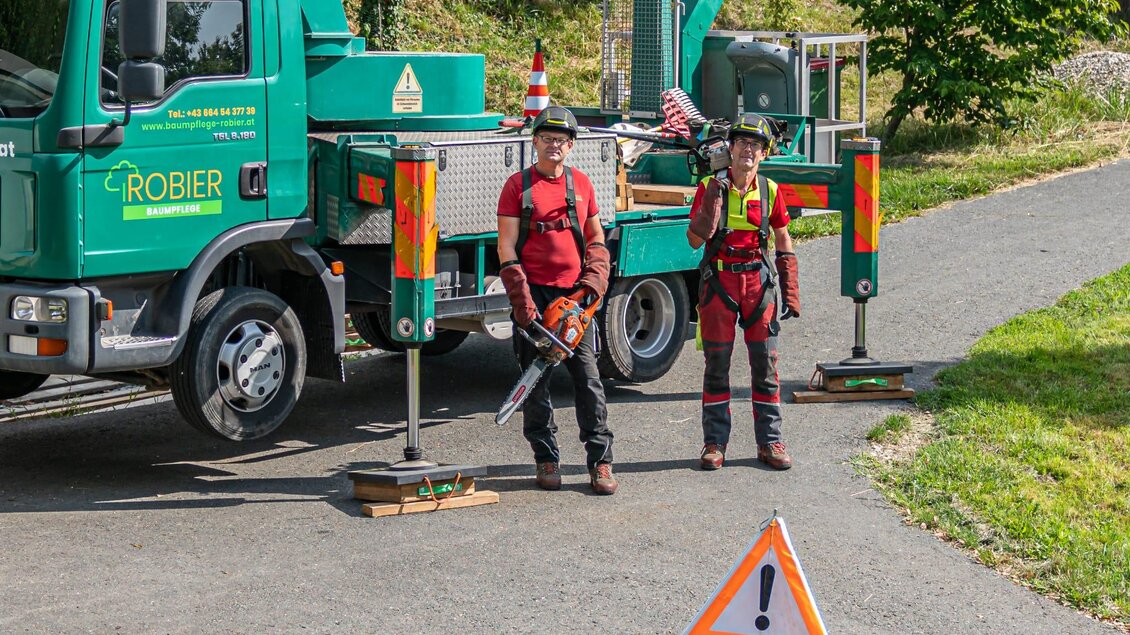 Zwei Personen in Arbeitskleidung stehen auf einem Weg neben einem Fahrzeug mit einem Baumdienst. Ein Warnschild weist auf Baumarbeiten hin. | © Baumpflege Robier