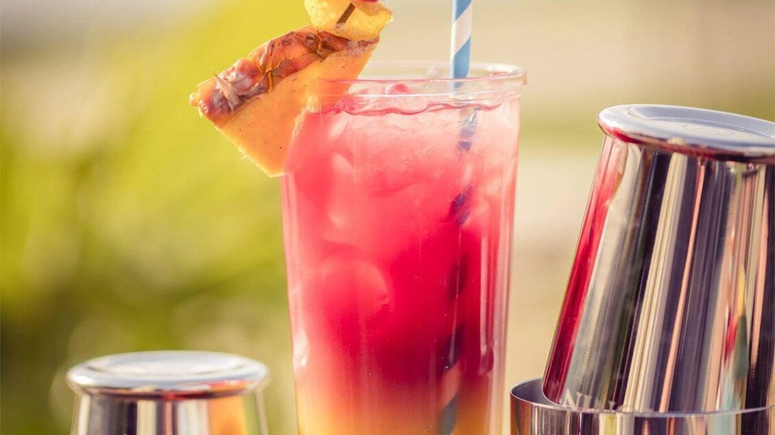 Ein bunter Cocktail mit einer Ananasscheibe, einer Himbeere und einem gestreiften Strohhalm. Der Drink steht vor glänzenden Barkeeper-Werkzeugen in einer sonnigen Umgebung. | © Beach Bar