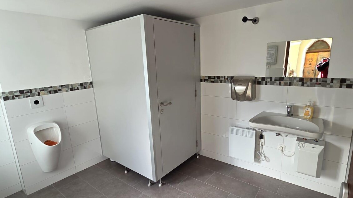 Ein modernes Badezimmer mit einer Duschkabine, einem Waschbecken und einem Urinal. Der Raum ist hell und sauber, mit grauen Fliesen und einer einfachen Ausstattung. | © Thomas Herzog
