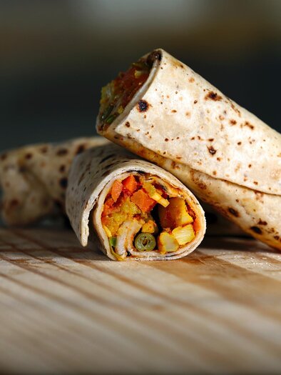 Ein aufgerollter Wrap mit buntem Gemüsefüllung liegt auf einem Holzbrett. Die Tortilla ist leicht braun und sieht frisch aus. | © Nishant Aneja