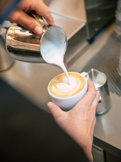 Eine Hand gießt geschäumte Milch in eine Tasse mit Kaffee. Der Kaffee hat eine schöne Latte-Art-Oberfläche. | © Bellaggio
