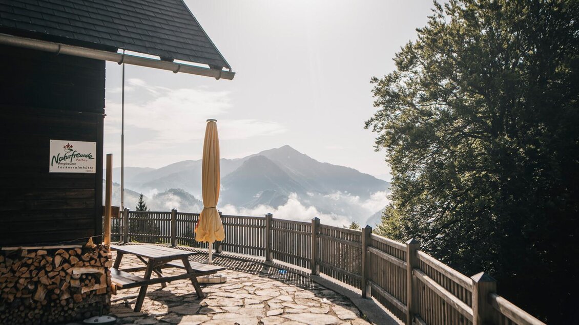 Ein gemütlicher Außenbereich mit einem Tisch und einer Sonnenschirm auf einem Steindeck. Im Hintergrund sind majestätische Berge und Nebel zu sehen. | © Stefan Leitner