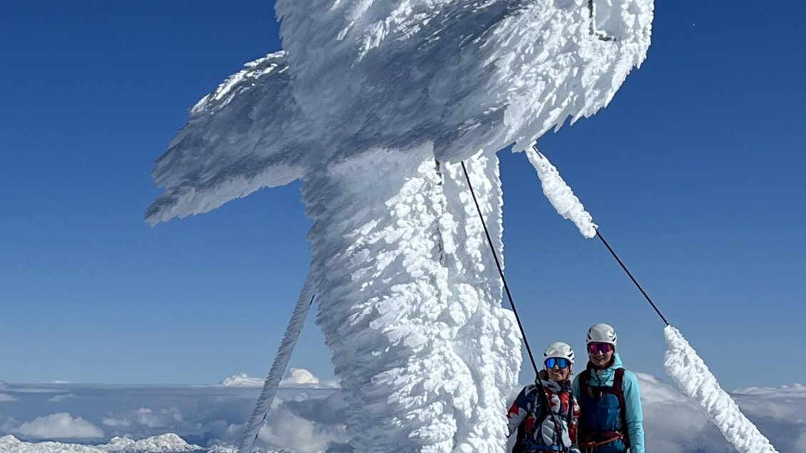 Ein schneebedeckter Berggipfel mit einem großen, gefrorenen Kreuz. Zwei Bergsteiger stehen vor dem Kreuz und genießen die Aussicht auf die umliegenden Berge. | © Bergführer Dachstein – Hans Prugger