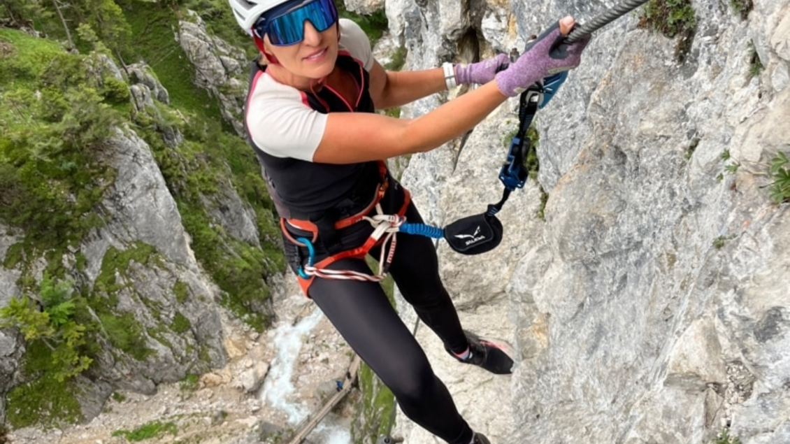 Eine Person klettert an einer steilen Felswand. Im Hintergrund sind bewaldete Berge und ein kleiner Fluss zu sehen. | © Hans Prugger