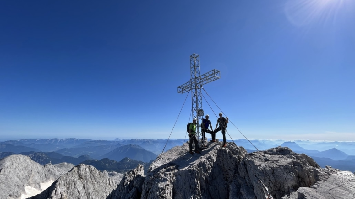 Eine Gruppe von Bergsteigern steht auf einem Gipfel mit einem Kreuz. Im Hintergrund erstrecken sich majestätische Berglandschaften und ein blauer Himmel. | © Hans Prugger