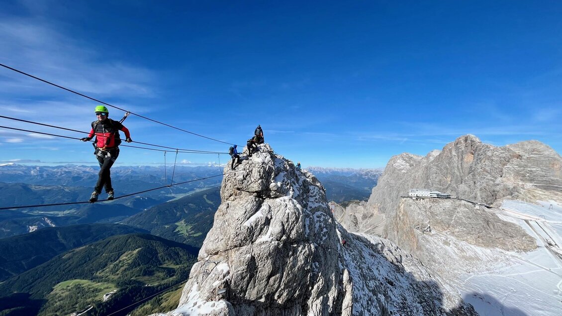 Ein Abenteurer läuft auf einer Ziplinestrasse über einen hohen Berggipfel. Die Landschaft zeigt beeindruckende alpine Ausblicke und einen klaren blauen Himmel. | © Hans Prugger
