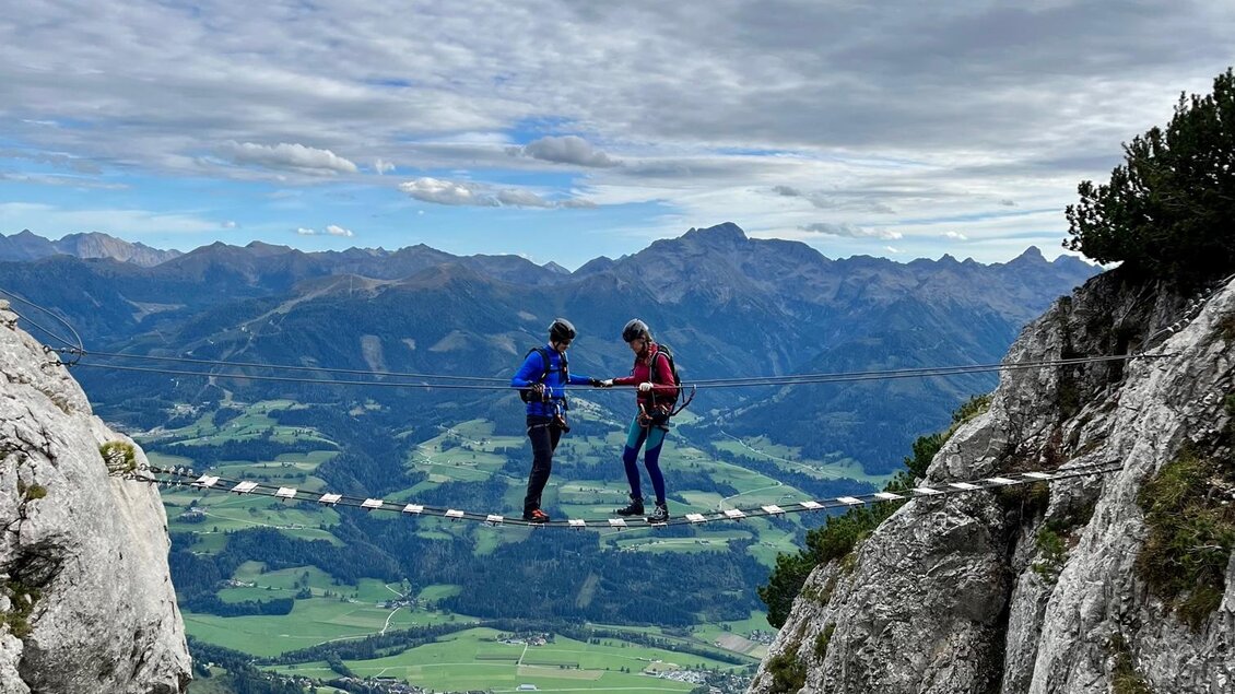 Zwei Kletterer stehen auf einem schmalen Grat zwischen Felsen. Im Hintergrund erstreckt sich eine grüne Landschaft mit Bergen und einem bewölkten Himmel. | © Hans Prugger