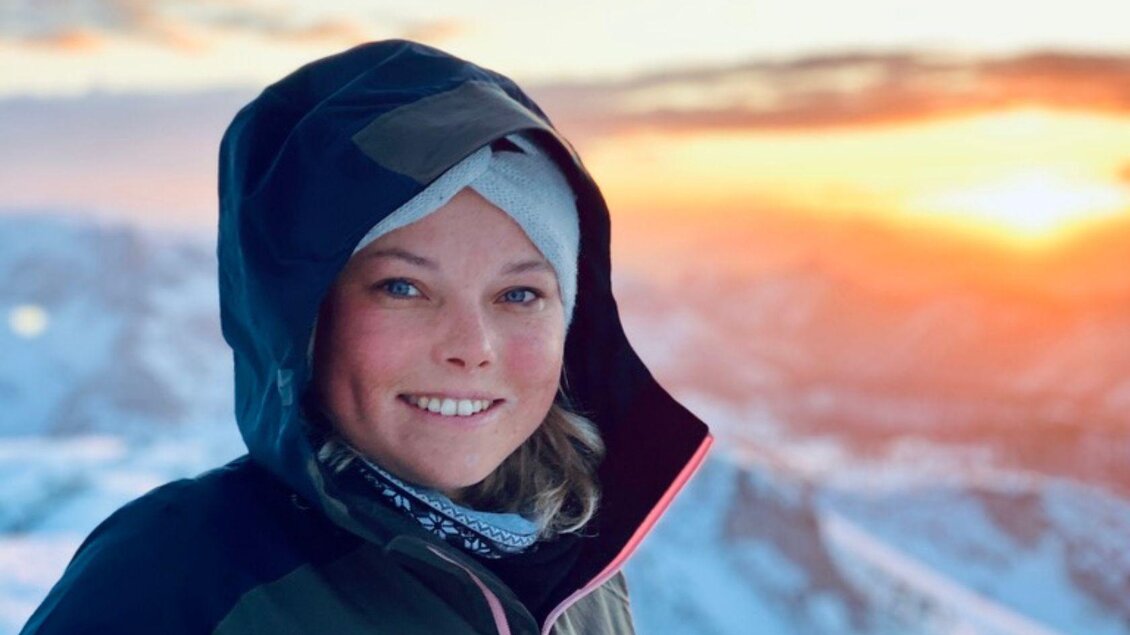 Eine junge, gutgelaunte Frau stahlt in die Kamera. Sie trägt eine Mütze und einen Anorak mit Kapuze, im Hintergrund erkennt man verschneite  Berggipfel in der untergehenden Sonne. | © Alice Hofbauer