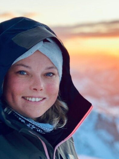 A young, cheerful woman smiles at the camera. She is wearing a cap and a hooded anorak, and in the background, you can see snow-covered mountain peaks in the setting sun. | © Alice Hofbauer