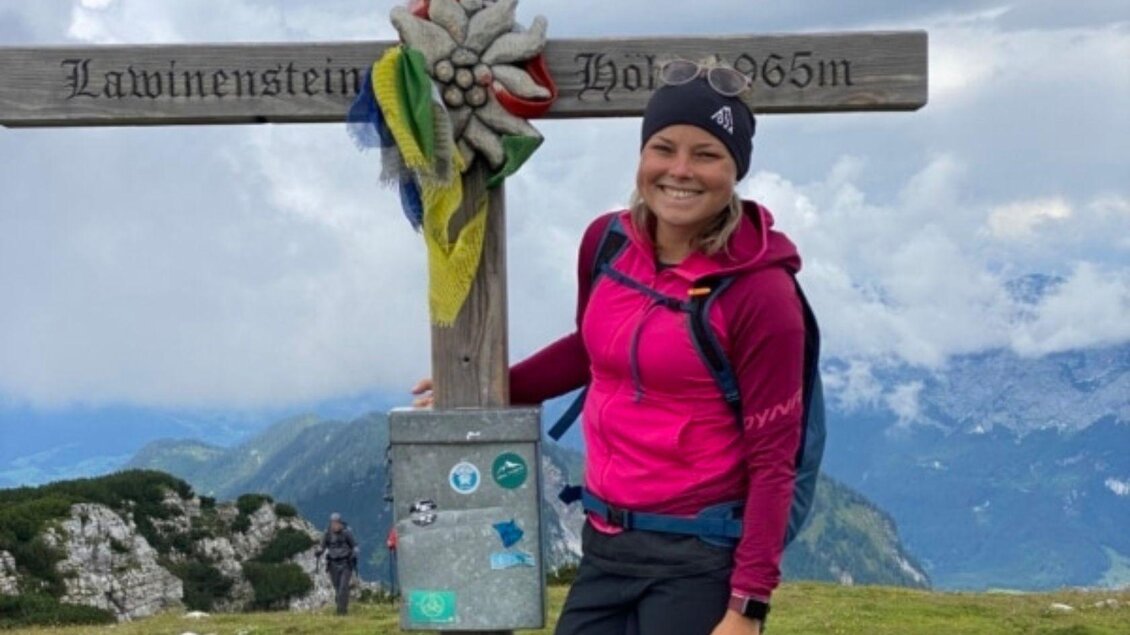Eine Junge, blonde Frau steht neben einem Gipfelkreuz und lacht in die Kamera. Sie trägt eine pinkfarbene Jacke und ein schwarzes Stirnband. | © Alice Hofbauer
