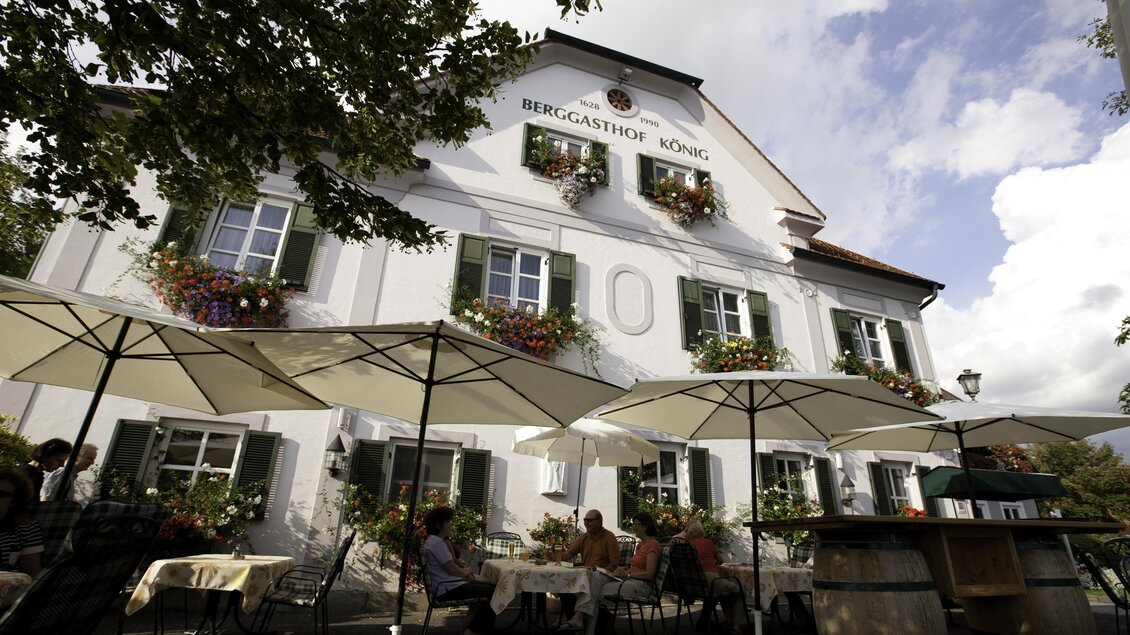 Ein gemütliches Gasthaus mit einer einladenden Terrasse. Tische mit gelben Tischdecken sind im Freien aufgestellt, umgeben von grünen Pflanzen. | © Berggasthof König