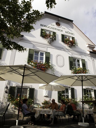 Ein gemütliches Gasthaus mit einer einladenden Terrasse. Tische mit gelben Tischdecken sind im Freien aufgestellt, umgeben von grünen Pflanzen. | © Berggasthof König