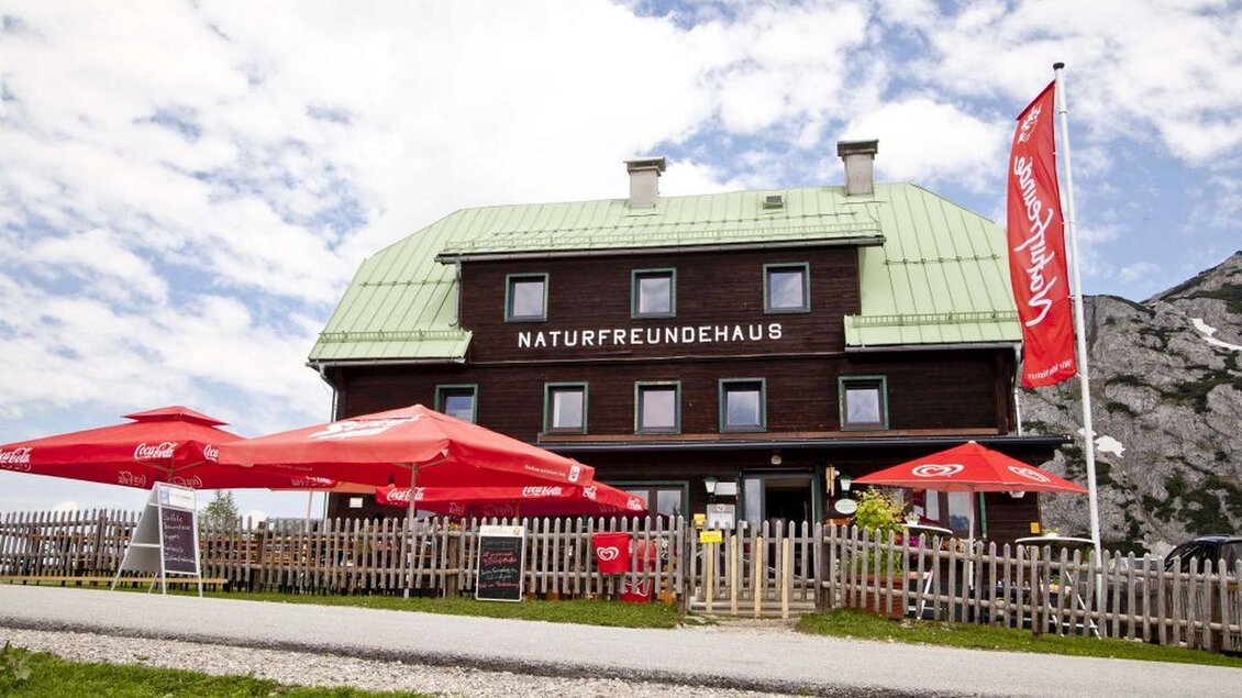 <p>Das gemütliches Naturfreundehaus mit einem grünen Dach und Terrassen unter roten Sonnenschirmen. Umgeben von einer malerischen Landschaft und montanem Ambiente.</p> | © Naturfreundehaus Tauplitzalm