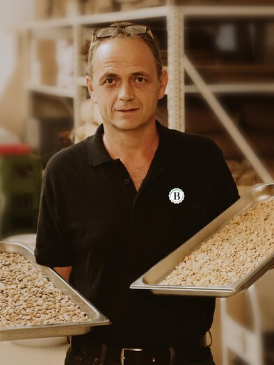 A man is holding two trays of nuts in his hand. The background shows a workshop with various materials. | © Berghofer Farmery