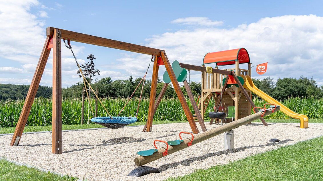 <p>Ein Spielplatz mit einer Schaukel, einer Wippe und einer Rutsche. Im Hintergrund sind grüne Felder und ein blauer Himmel zu sehen. Bergschenke Schlögl in Kirchberg an der Raab</p> | © Andreas Schlögl