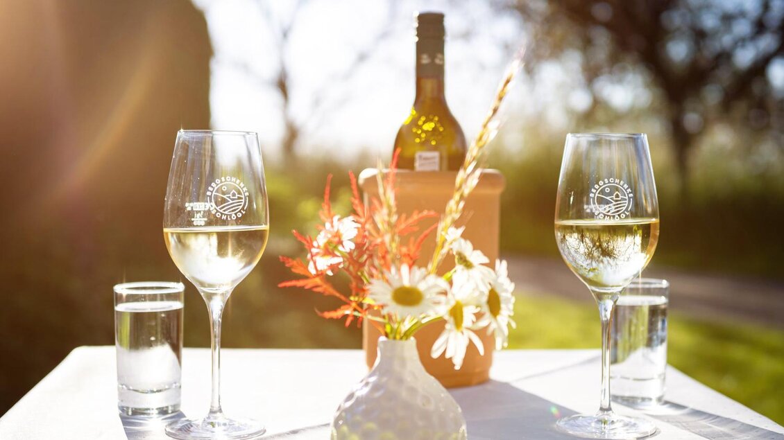 <p>Zwei Weingläser stehen auf einem Tisch neben einer Vase mit Blumen. Im Hintergrund ist eine Flasche Wein zu sehen, und die Sonne scheint hell. Bergschenke Schlögl in Kirchberg an der Raab</p> | © Andreas Schlögl