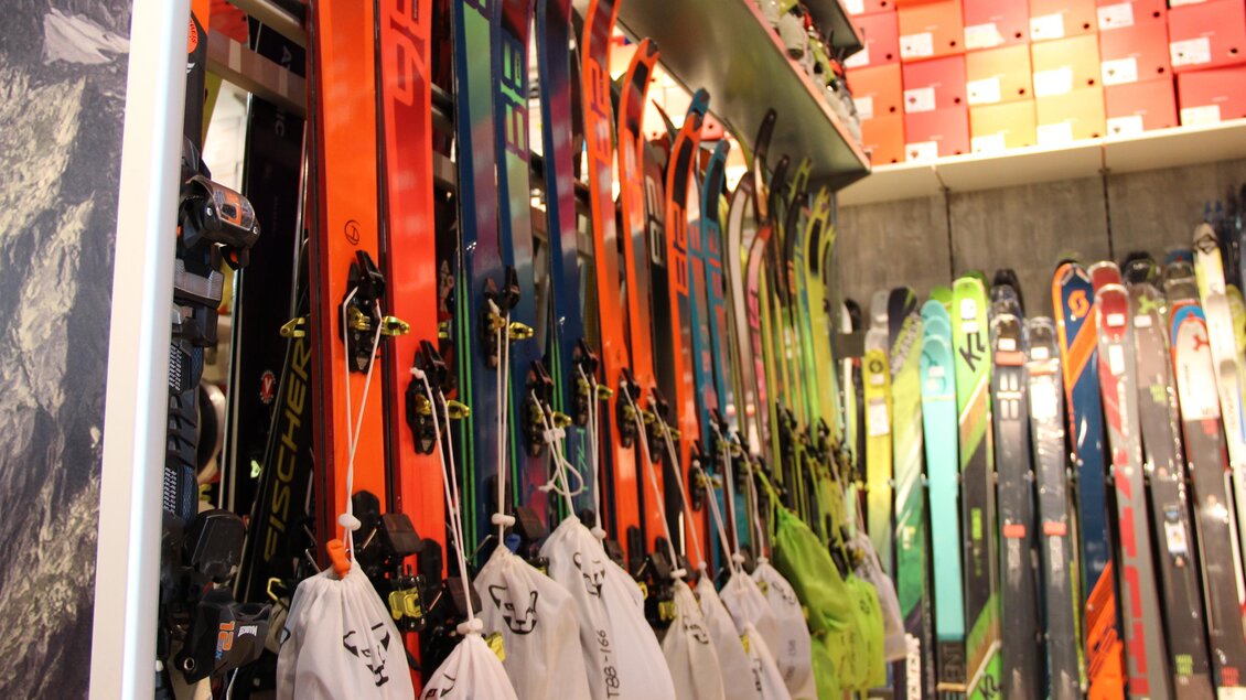 Ein Skishop mit vielen Skiern in verschiedenen Farben ordentlich aufgereiht. In der Ecke sind weiße Skitaschen sichtbar.