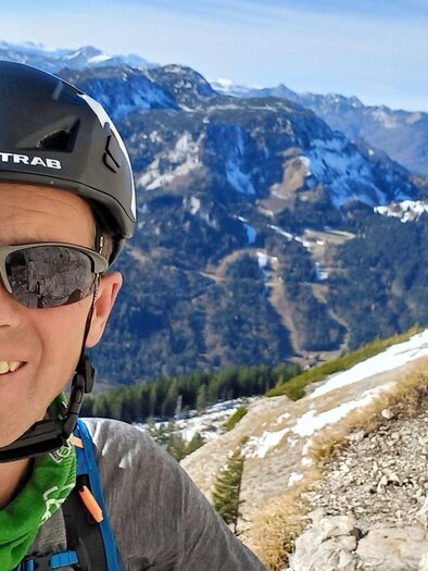 A mountaineer with a helmet and sunglasses poses in front of an impressive mountain landscape. The view shows high mountains and a clear blue sky. | © Gerhard Ziermayer