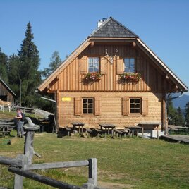Holzhütte Bernsteinhütte in Hirschegg-Pack mit Blumenkästen, Sitzplätzen und Wanderweg in alpiner Landschaft. | © TV Lipizzanerheimat | EU