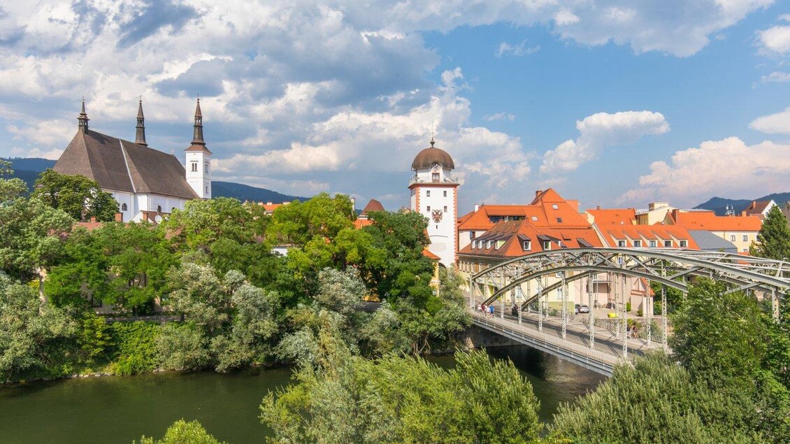 Eine malerische Stadtansicht mit einer alten Kirche und einem historischen Turm. Eine Brücke führt über einen ruhigen Fluss, umgeben von Bäumen und Häusern. | © TV ERZBERG LEOBEN