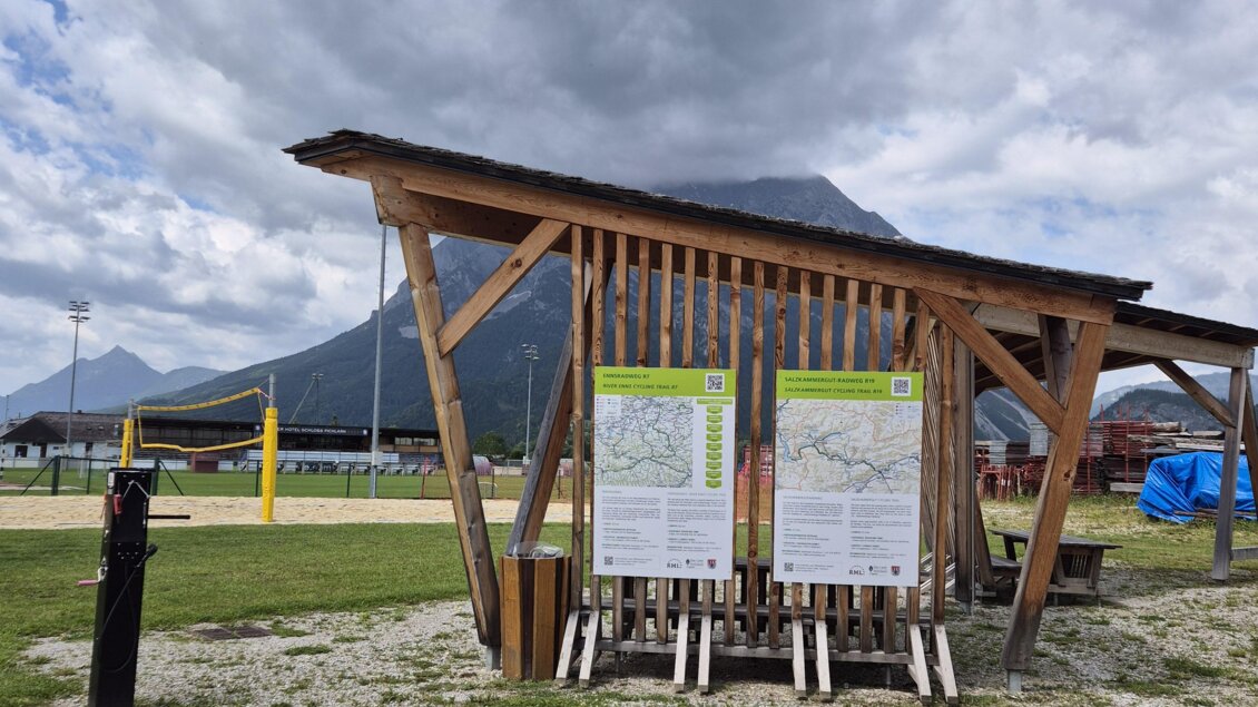 Ein Informationsstand aus Holz steht auf einem Platz mit Blick auf die Berge. Der Himmel ist bewölkt und es gibt viel Grün um den Stand herum. | © TVB Schladming-Dachstein