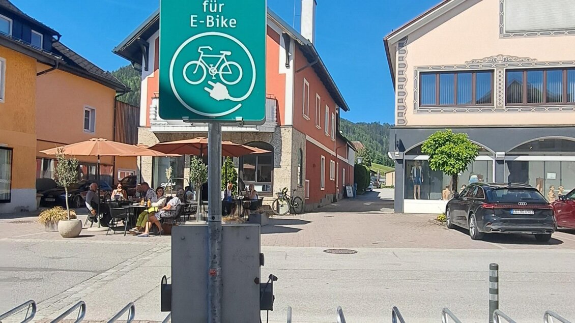 Ein Radweg-Schild steht an einer Straße mit Häusern im Hintergrund. Auf der linken Seite sind Fahrradständer und ein Restaurantbereich sichtbar.