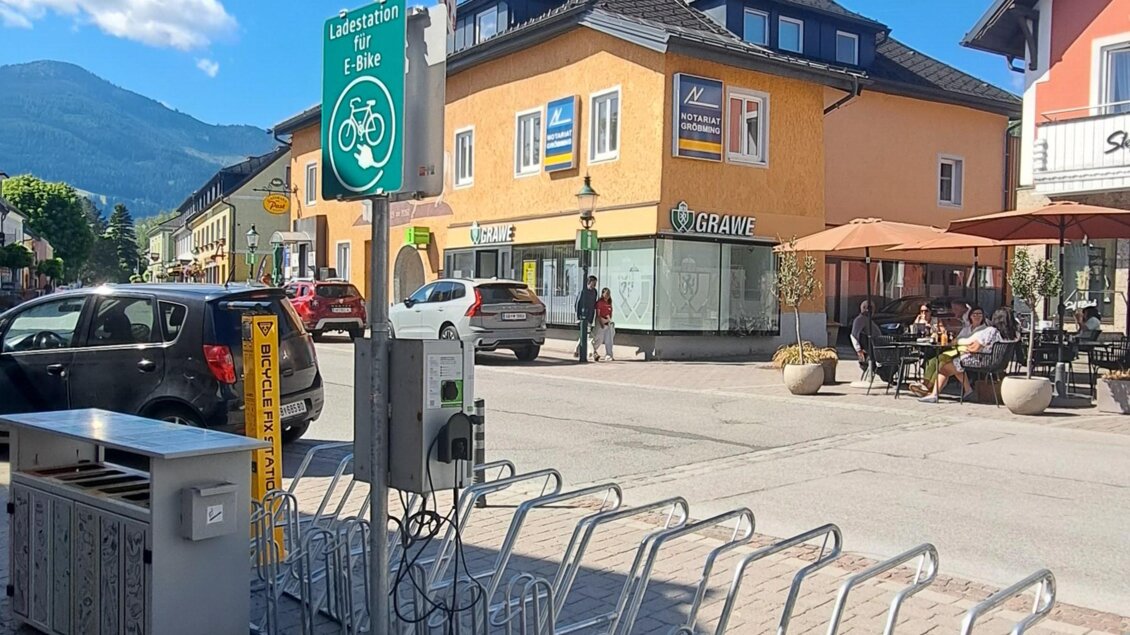 Eine Straßenansicht mit einem klaren blauen Himmel und einem bewachsenen Platz. Zu sehen sind orangefarbene Gebäude und Fahrradständer.