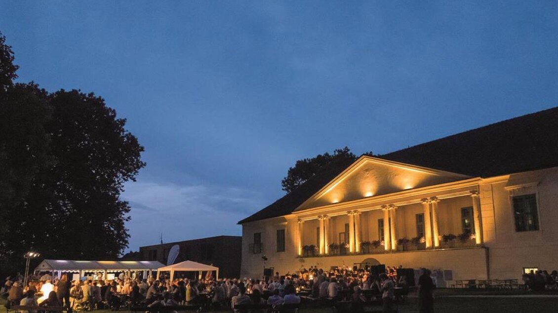 Eine festliche Veranstaltung im Freien mit vielen Gästen vor einem beleuchteten Gebäude. Der Abendhimmel ist dunkelblau und es gibt gemütliche Sitzgelegenheiten. | © Michael Baumann