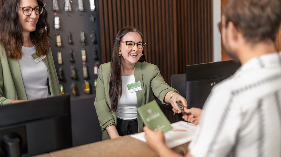 Zwei freundliche Hotelmitarbeiterinnen stehen am Empfang und lächeln. Eine von ihnen überreicht einem Gast einen Zimmerschlüssel.