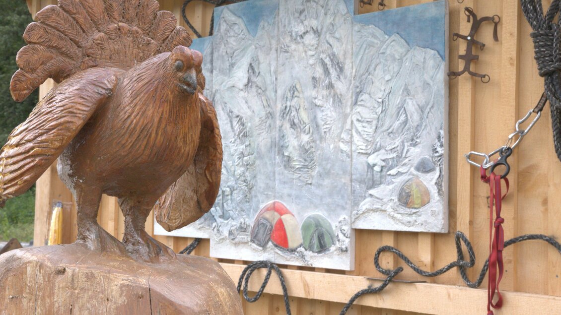 Eine Holzskulptur eines Vogels steht neben einer Wand mit einem Gemälde von Bergen und Zelten. Der Hintergrund besteht aus Holz mit verschiedenen Ketten und Seilen. | © Josef Schiestl